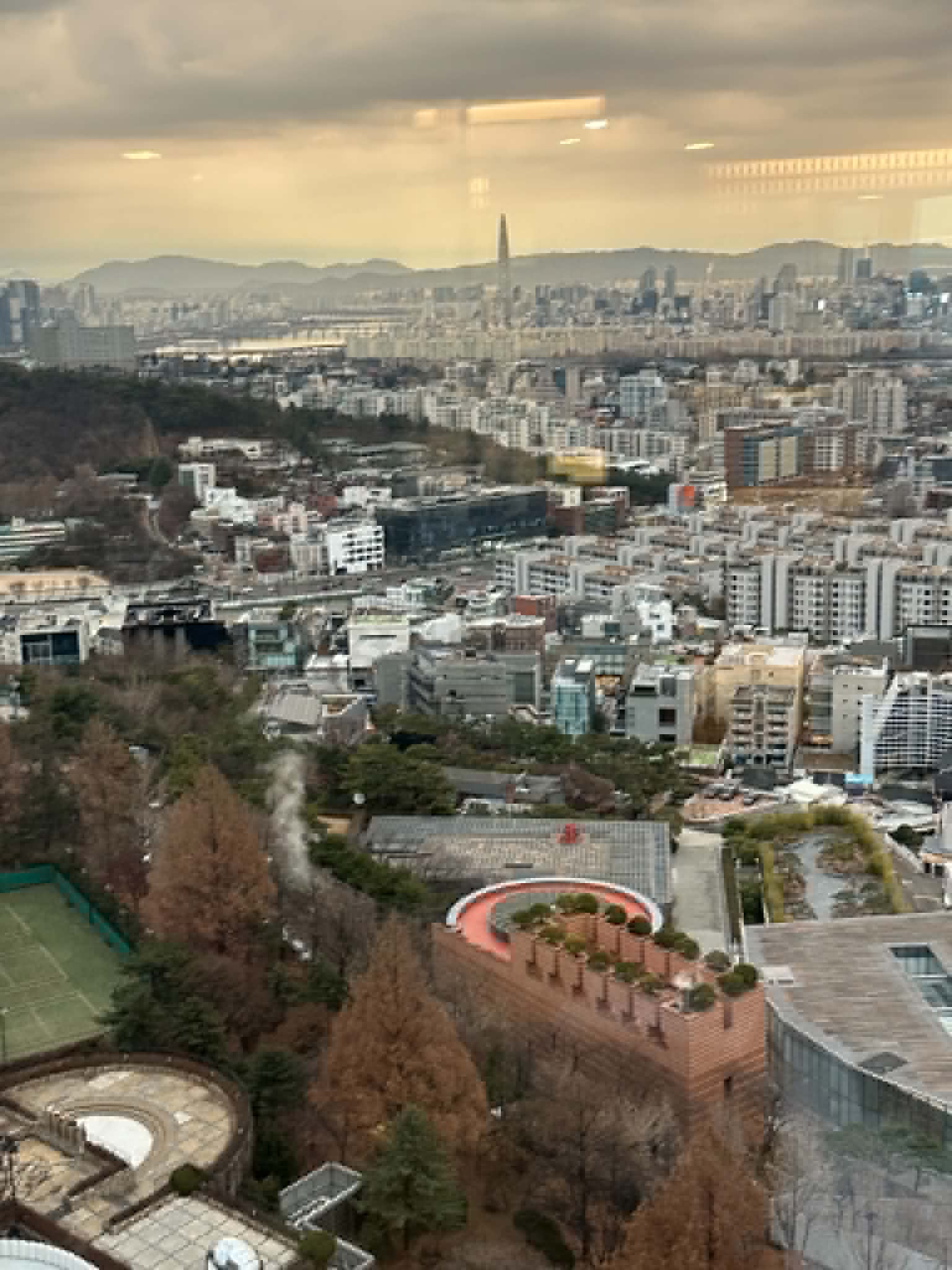 Seoul cityscape at dusk from a hotel high-floor, with Lotte World Tower in the distance and a tennis court in the foreground
