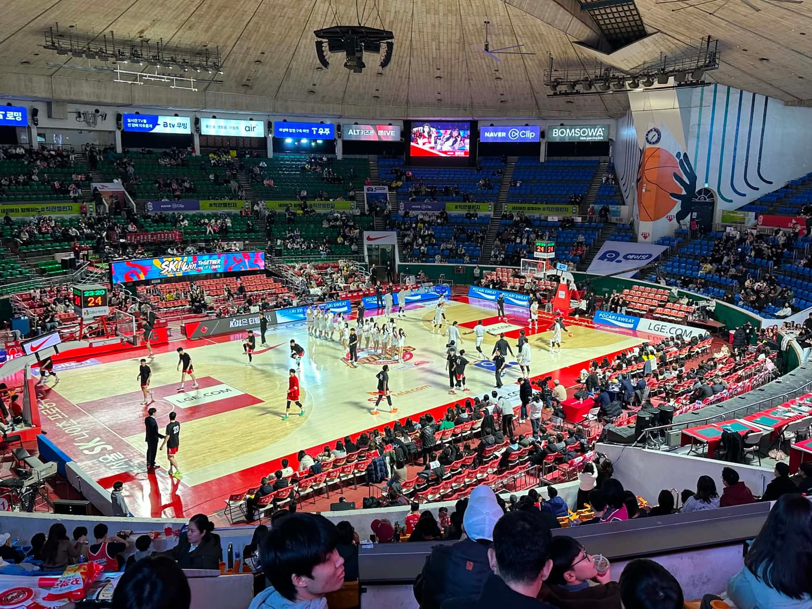 Wide shot of a Seoul SK Knights basketball arena with substantial crowd, players warming up, and Korean advertising visible