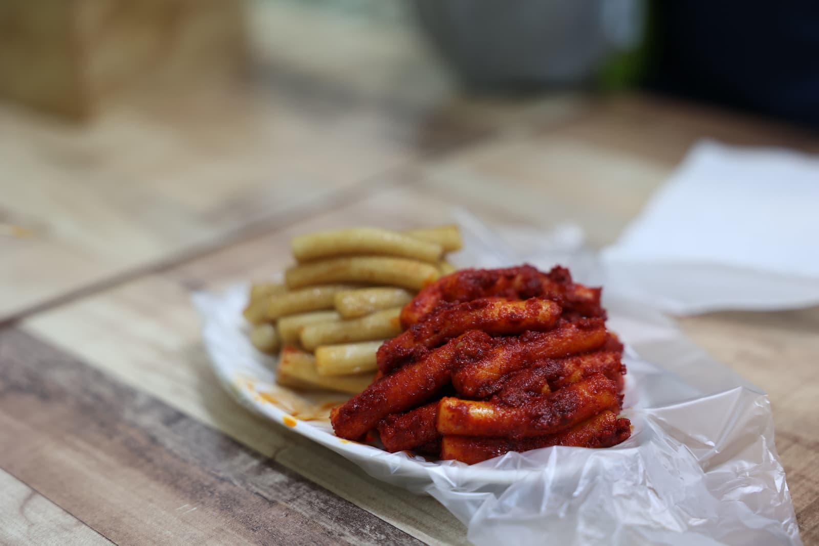 Plate of tteokbokki — Korean rice cakes — half coated in red gochujang sauce and half plain golden, on paper wrapping
