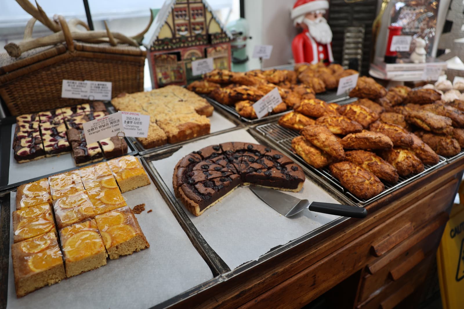Korean bakery counter with Christmas decorations — Santa figure, gingerbread house, antlers — and pastries including a chocolate tart and orange vanilla cake