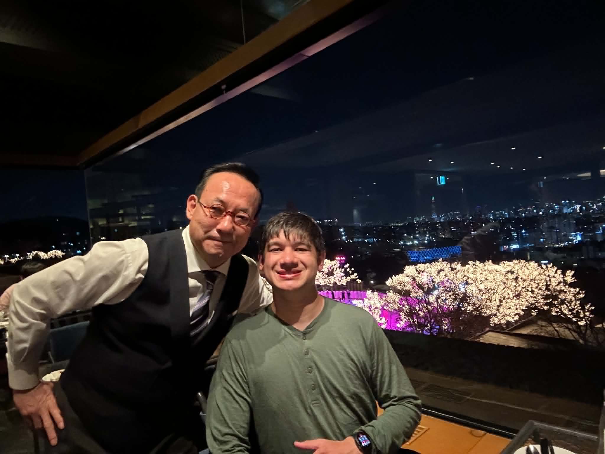 Selfie of Tommy Lee in a vest and tie next to Anthony in a green henley, with Seoul night skyline and lit trees through the restaurant window behind