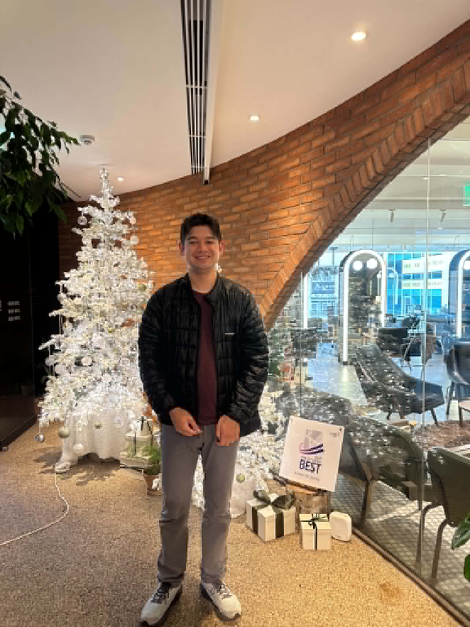 Anthony at Juno salon in a black puffer jacket, smiling, with a white Christmas tree to the left and a brick arch behind