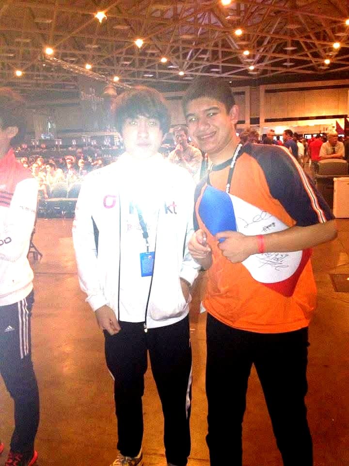 Teen Anthony in an orange jersey standing next to Korean StarCraft pro Flash in his white KT Rolster jacket at MLG Dallas, holding a signed flag