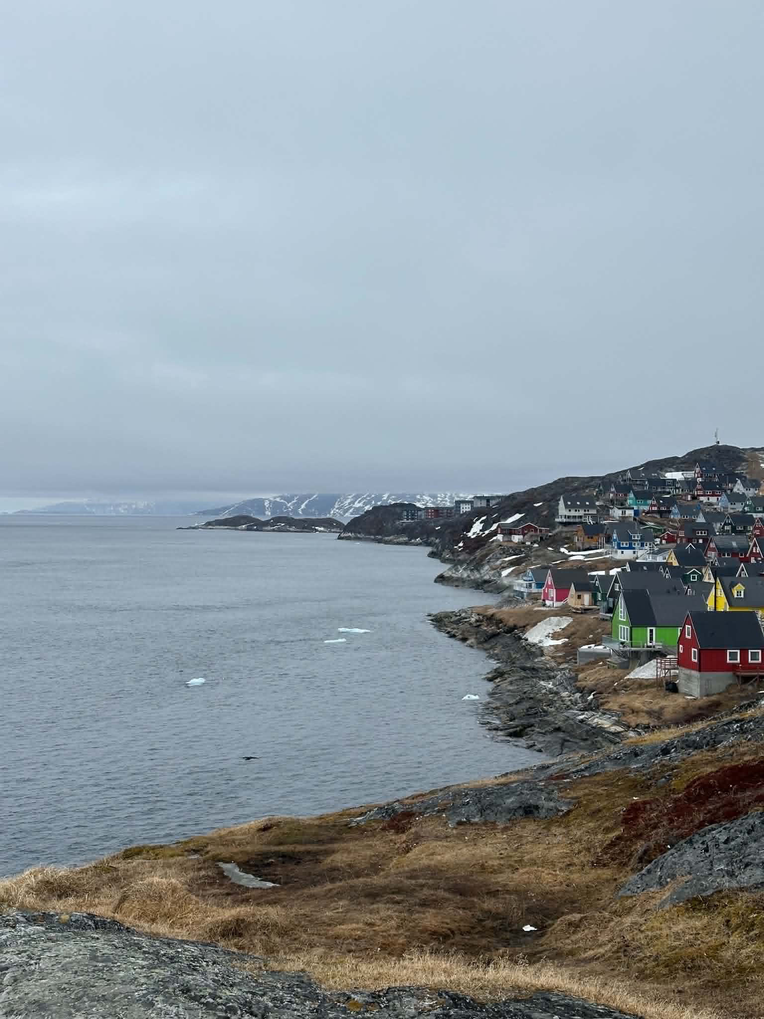 Pastel houses tumbling down a rocky coastline in Nuuk, with snow-capped mountains across the bay