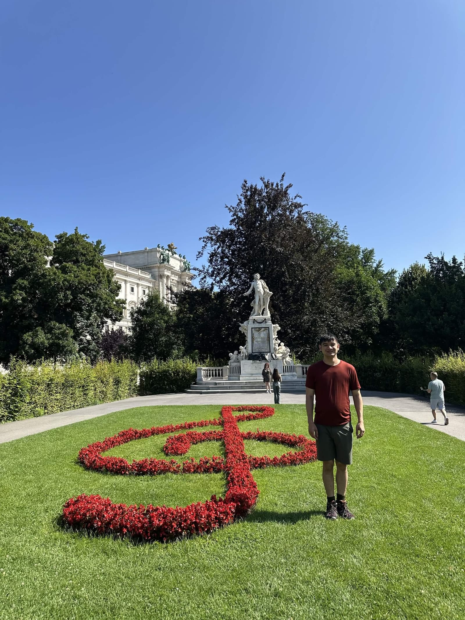 Anthony in a maroon t-shirt on the lawn of the Burggarten beside a large treble clef planted in red flowers, with the white marble Mozart monument and the Hofburg behind under a clear blue sky