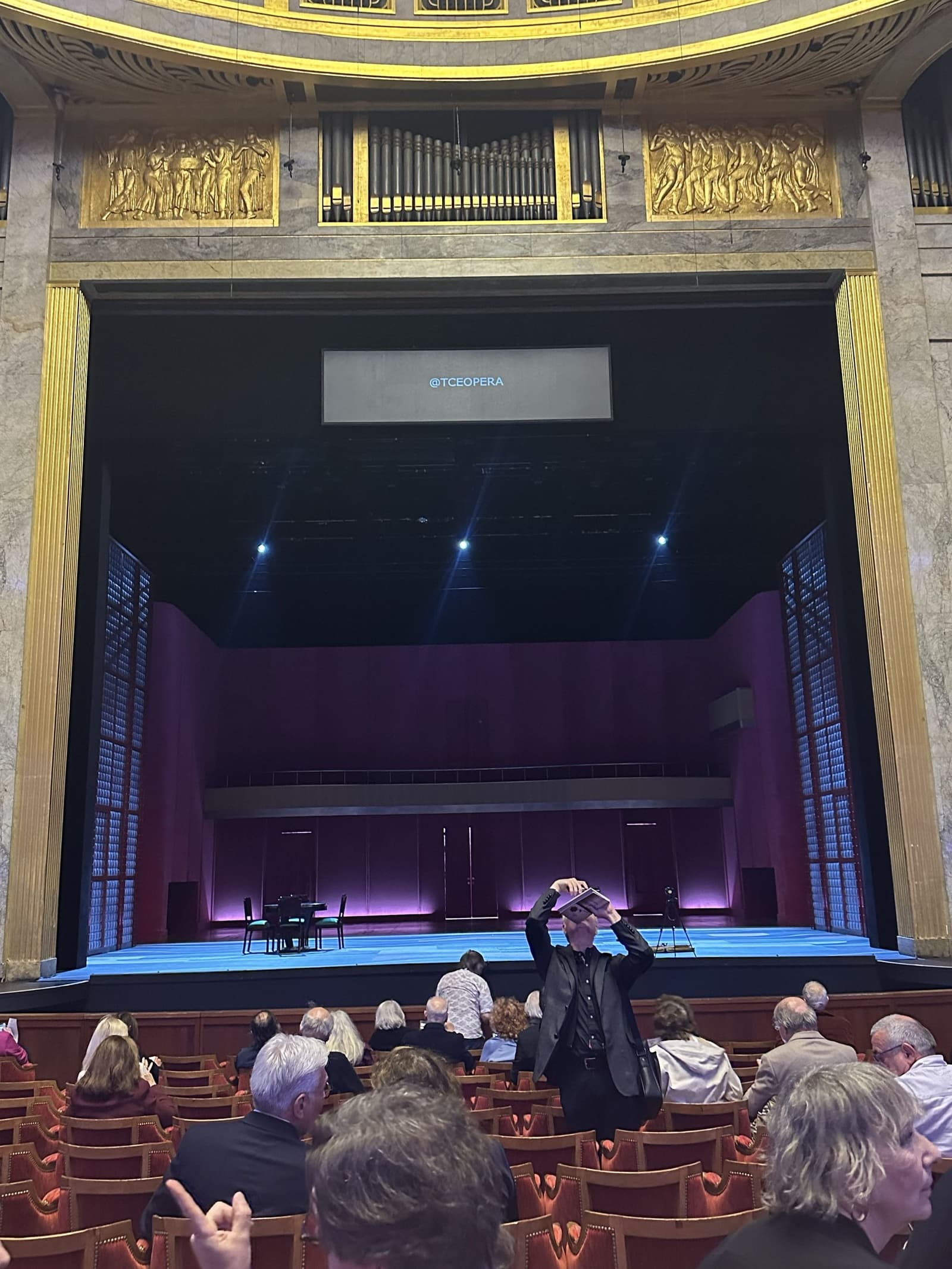 The stage of the Théâtre des Champs-Élysées set for a contemporary Der Rosenkavalier — a sparse purple-and-blue modern set with a small table and chairs, a screen reading @TCEOPERA above, and the audience seated below