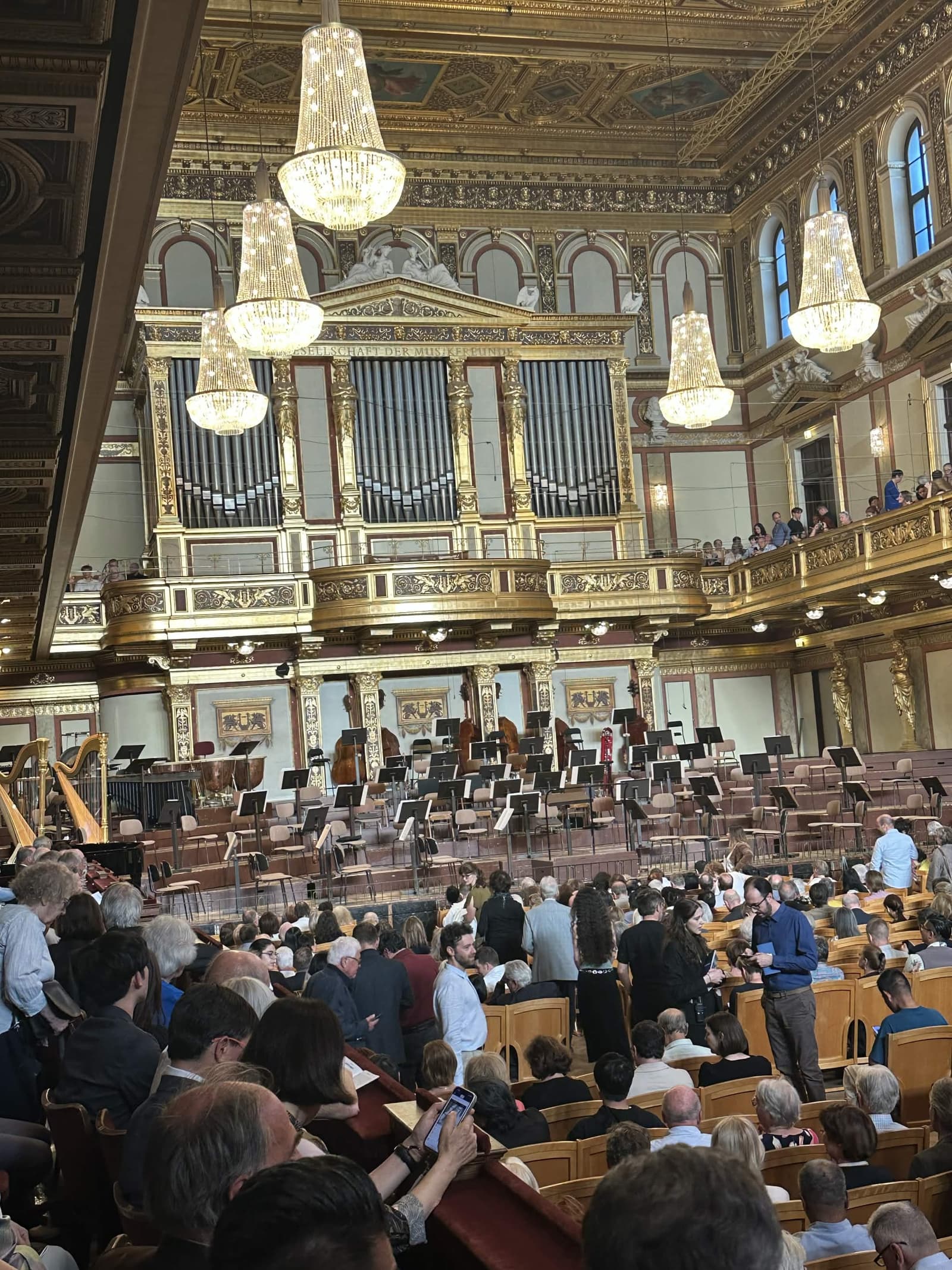 The gilded Golden Hall of the Vienna Musikverein before a concert — crystal chandeliers, gold balconies, a pipe organ, empty orchestra chairs and music stands on stage, and the audience filing in